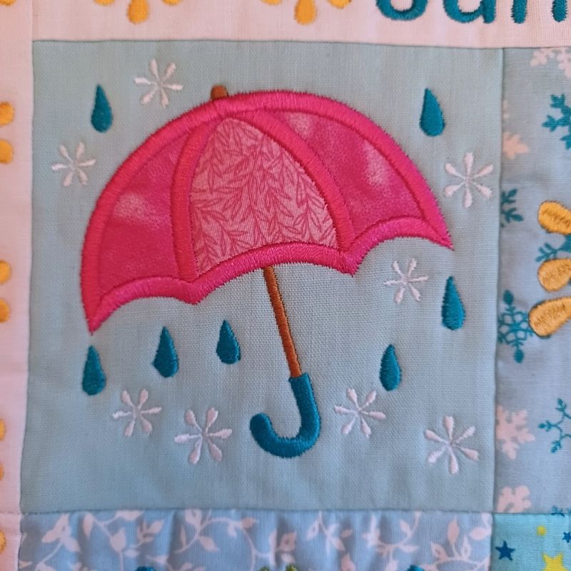 Close-up of ITH mini house quilt June embroidery design showing  appliqué  pink umbrella with raindrops on a fabric background
