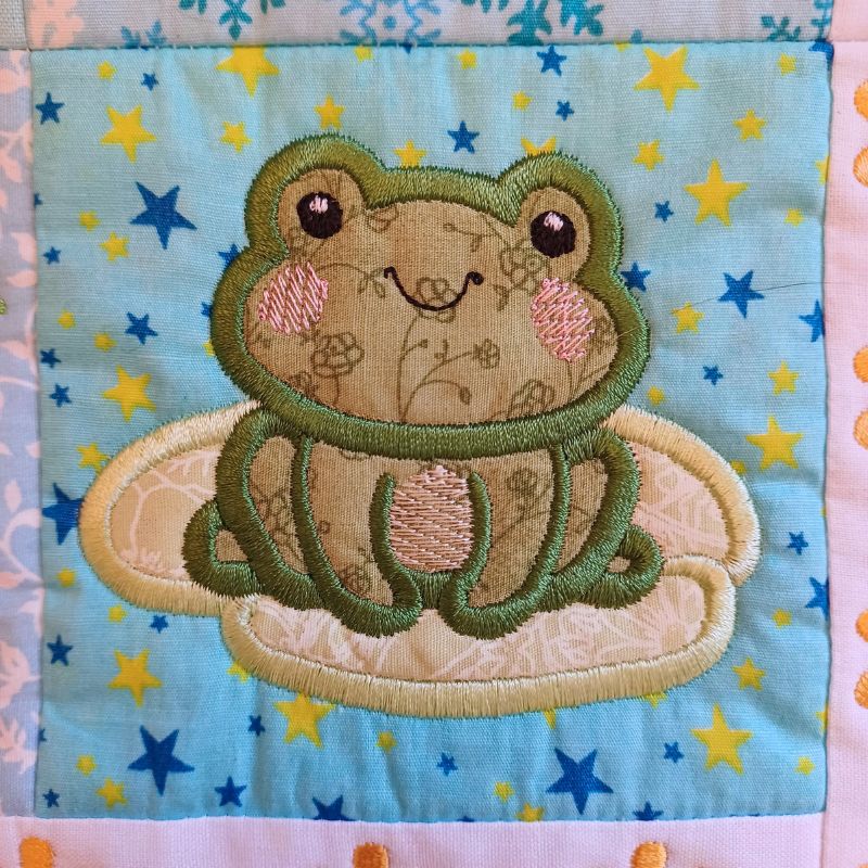 Close-up of ITH mini house quilt June embroidery design showing  appliqué frog design on a fabric background with star patterns