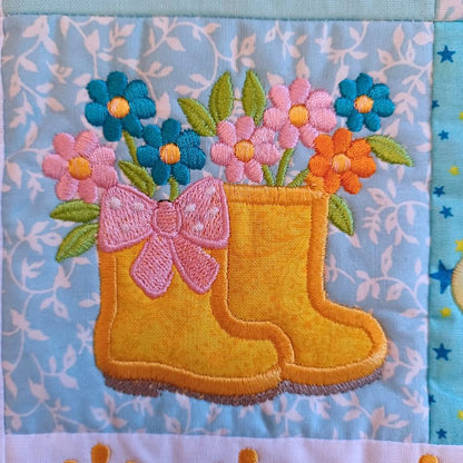 Close-up of ITH mini house quilt June embroidery design showing  appliqué yellow rain boots with pink bow and flowers on a light blue background